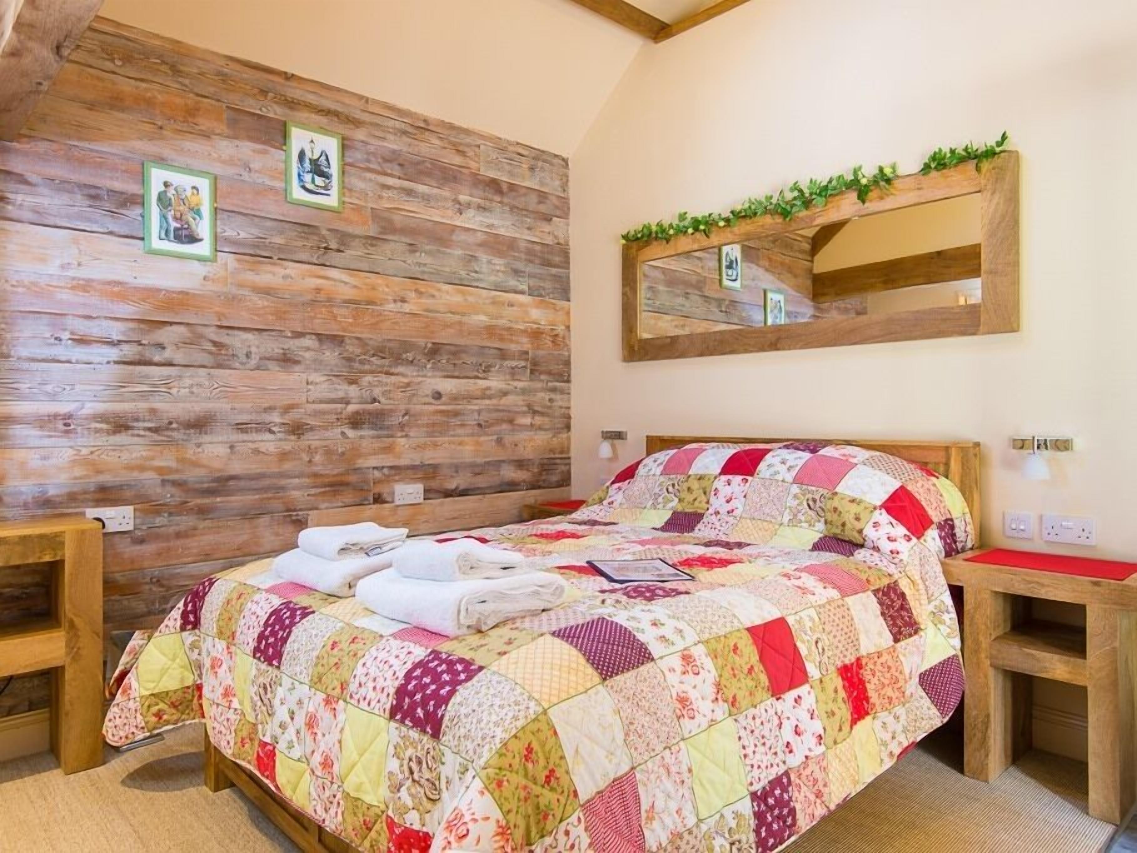Un dormitorio con pared de troncos de madera, colcha acolchada, mesita de noche de madera, espejo y una pequeña estantería con plantas.