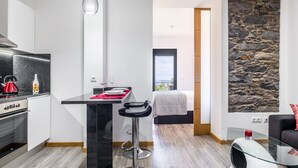 A modern kitchen with a black countertop, a red kettle, and a wine bottle. There is a dining area with a table set for two, a black chair, and a white cat bed. The room has a stone wall and a view of the outdoors.