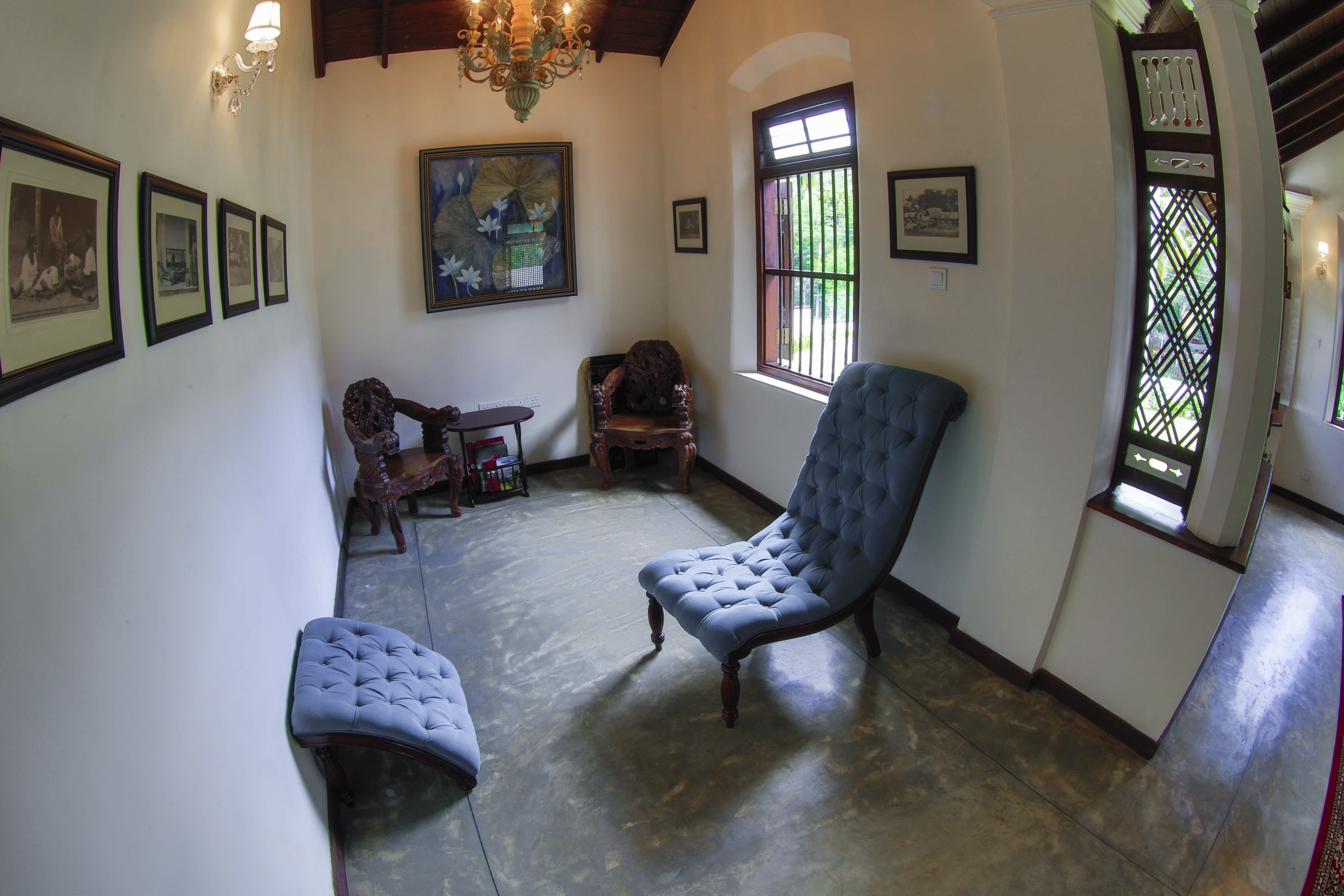 A spacious room with a large blue armchair, a smaller blue bench, and a wooden table with chairs. There are framed pictures on the walls and a chandelier.