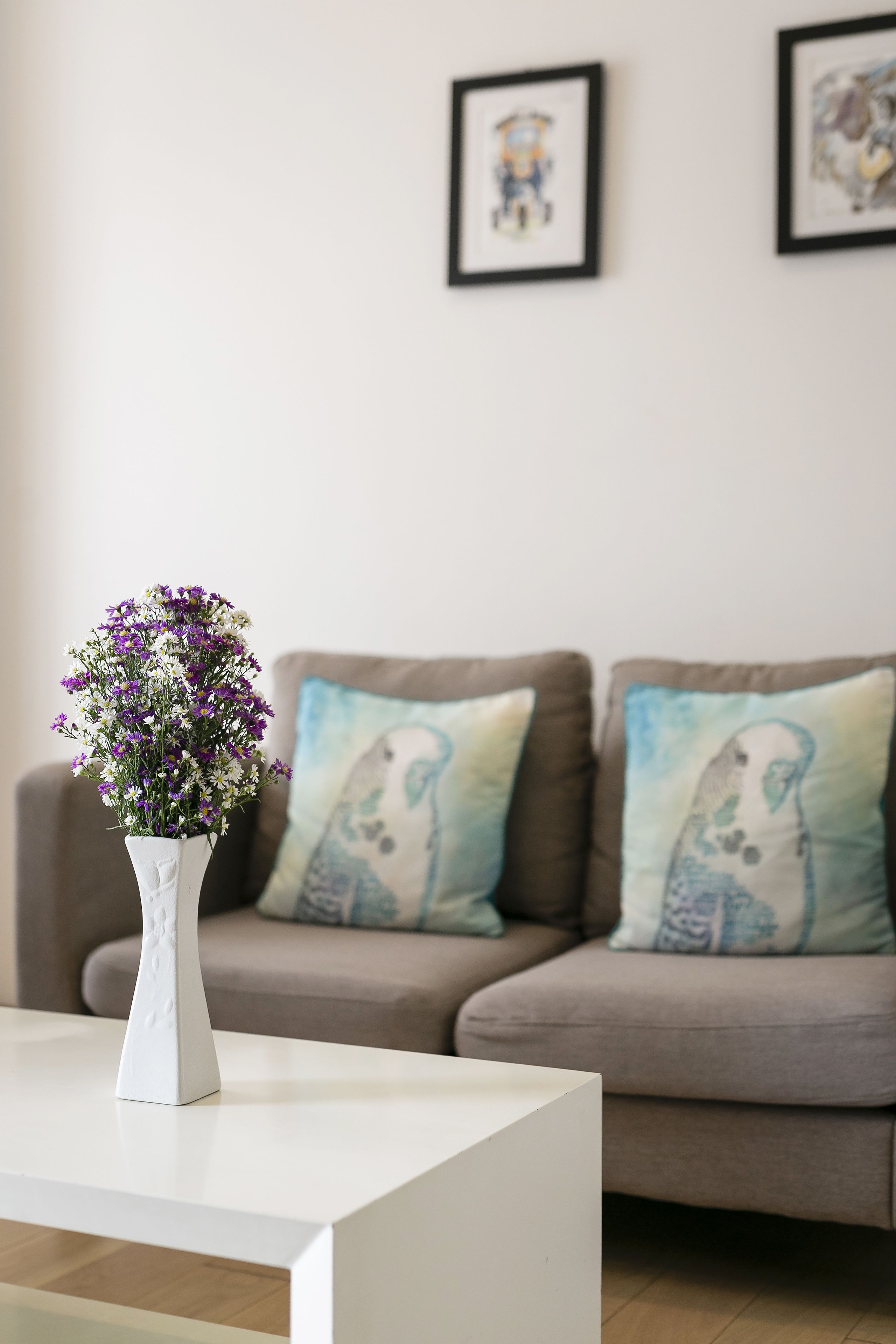 A white vase with purple flowers on a white table in a living room with a brown sofa and blue and white pillows.