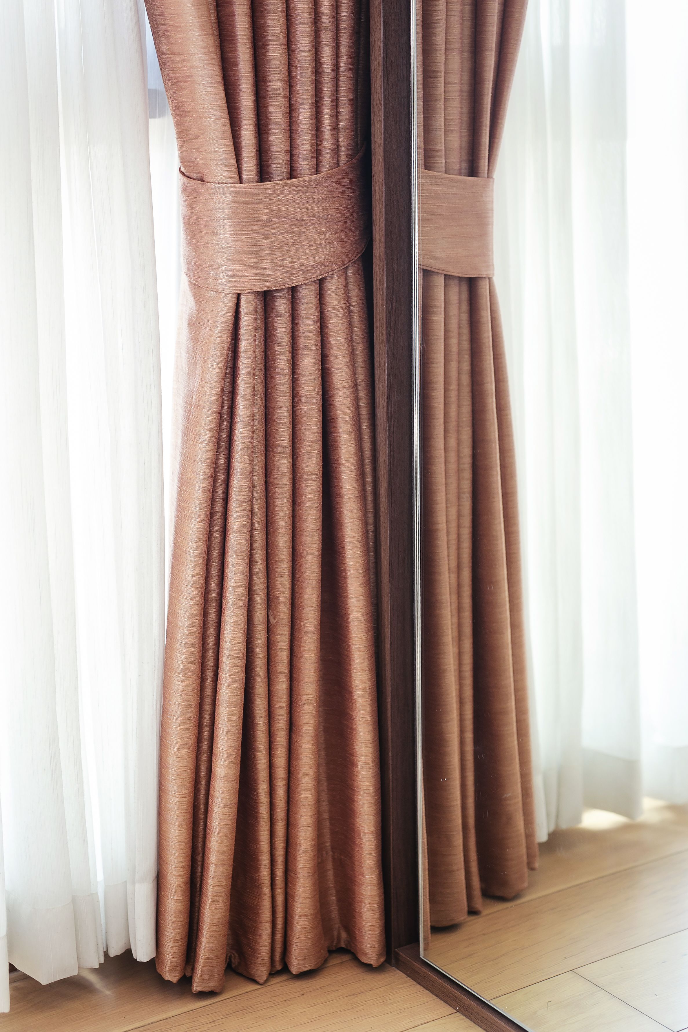 A window with brown curtains tied back, revealing a view of a wooden floor.