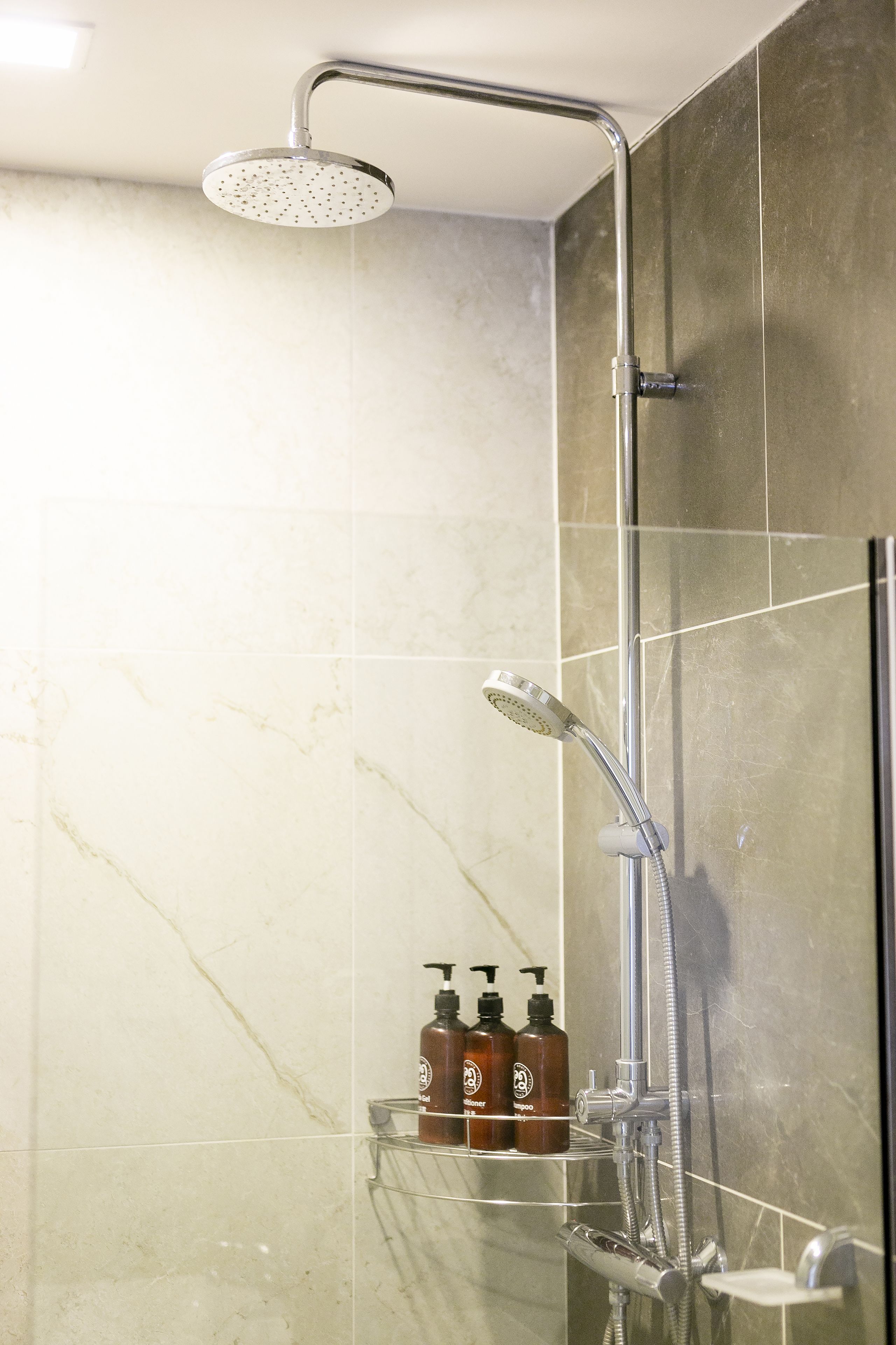 A modern shower with a handheld showerhead and a wall-mounted faucet. There are three bottles of shampoo on a shelf below.