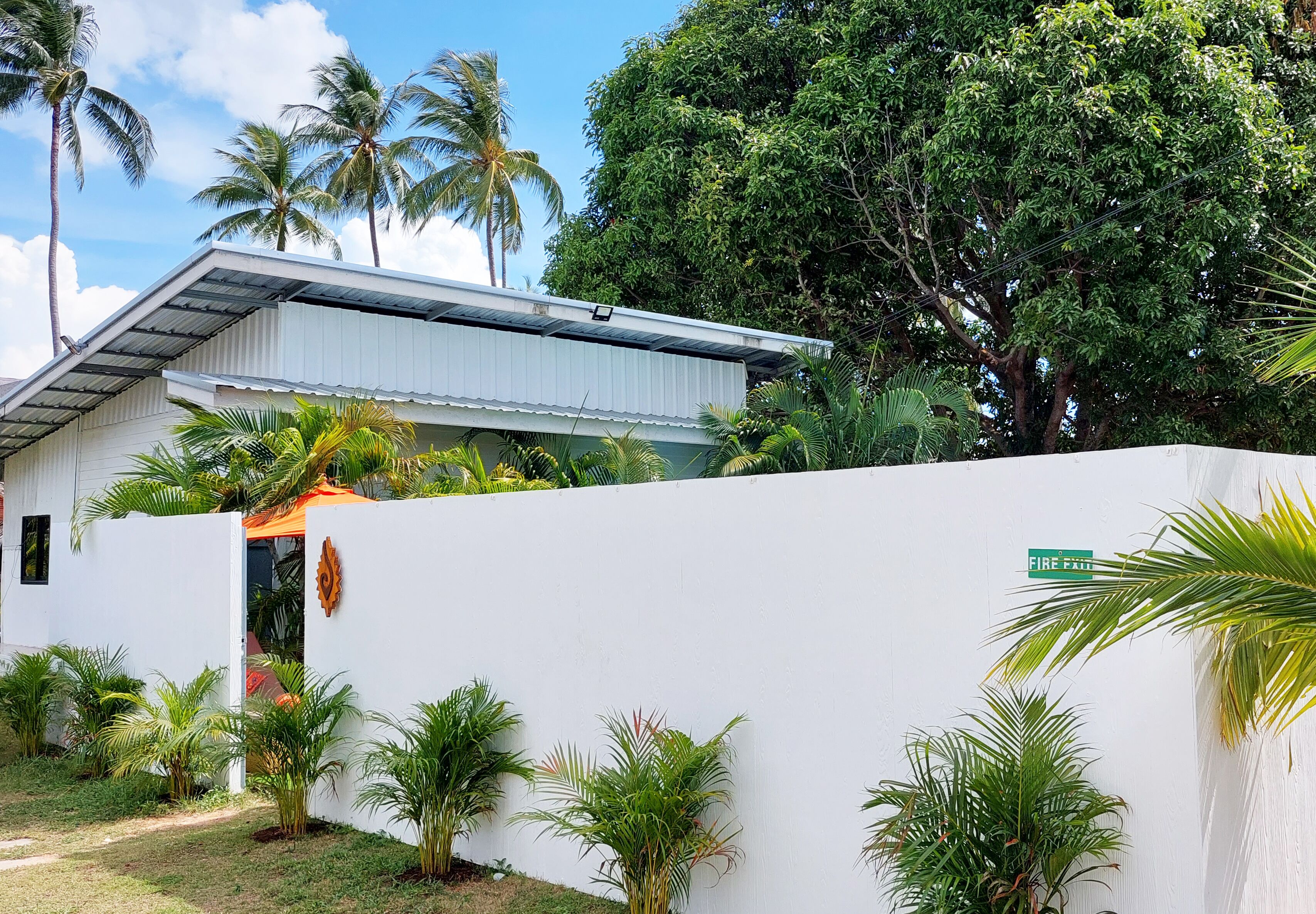 Un edificio blanco con techo metálico, rodeado de abundante vegetación y palmeras.