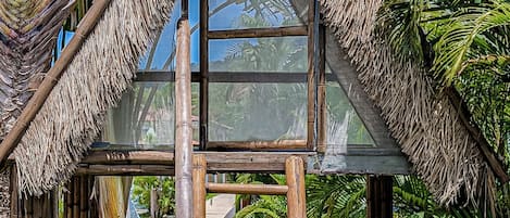 A thatched-roof structure with a ladder leading to a glass window, surrounded by lush greenery.