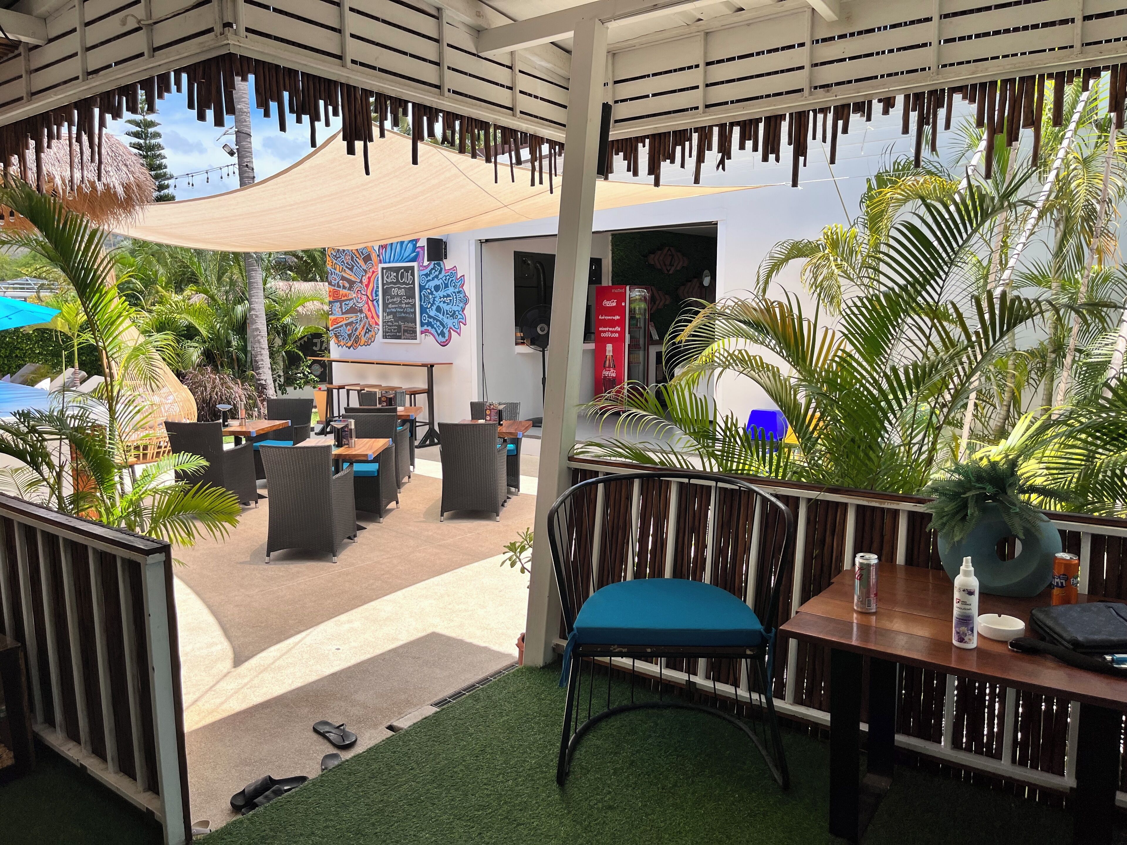 An outdoor seating area with tables and chairs, surrounded by greenery and a thatched roof structure.