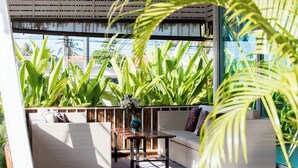 A sunlit outdoor seating area with a wooden table and white wicker sofas, surrounded by lush green plants. - Maya Resort (Koh Samui)