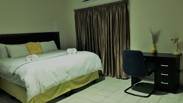 A hotel room with a bed, a desk, a chair, an air conditioner, and a curtain. - Essence of Africa Guesthouse (Windhoek)