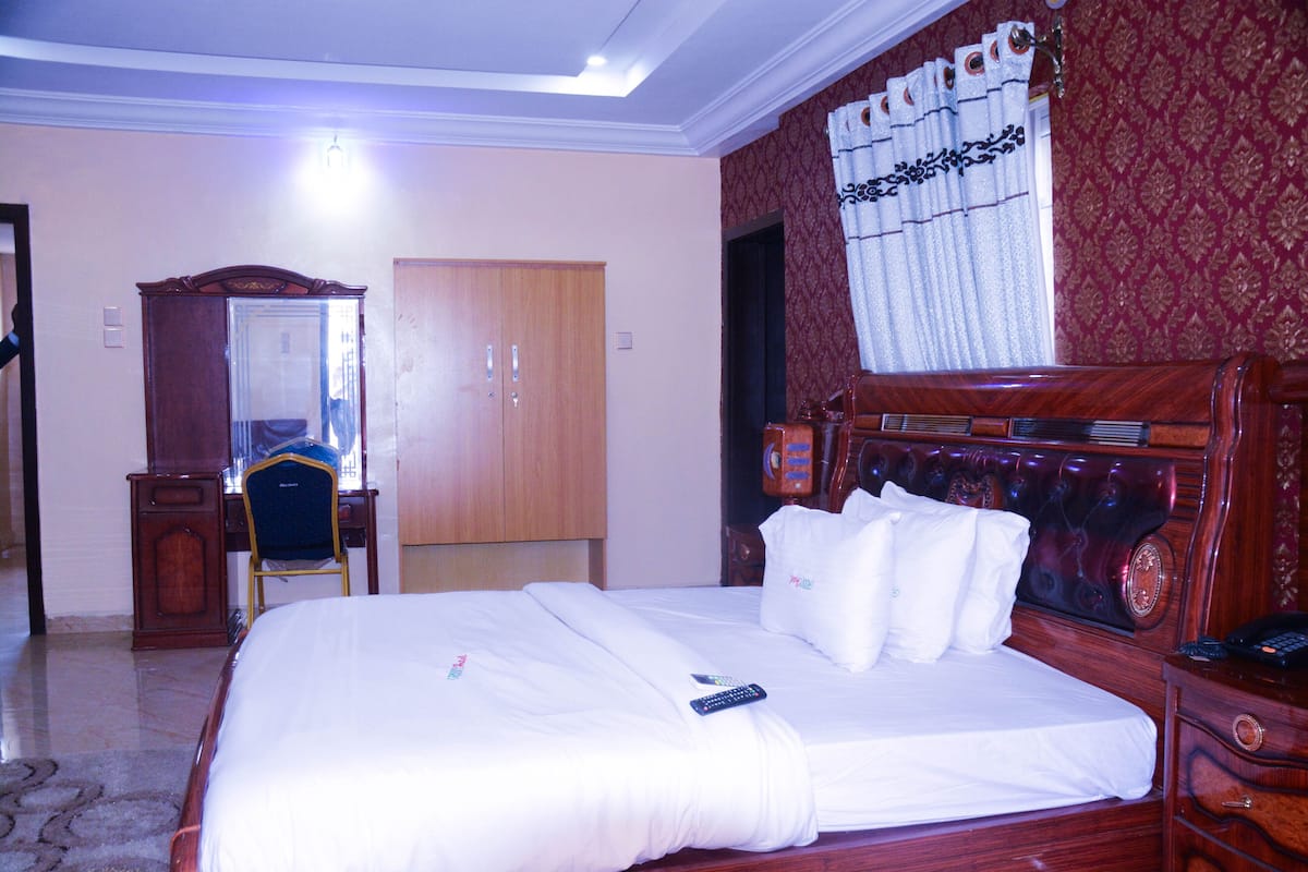 Quarto de hotel com uma cama grande, cabeceira de madeira, escrivaninha com cadeira, guarda-roupa e espelho.
