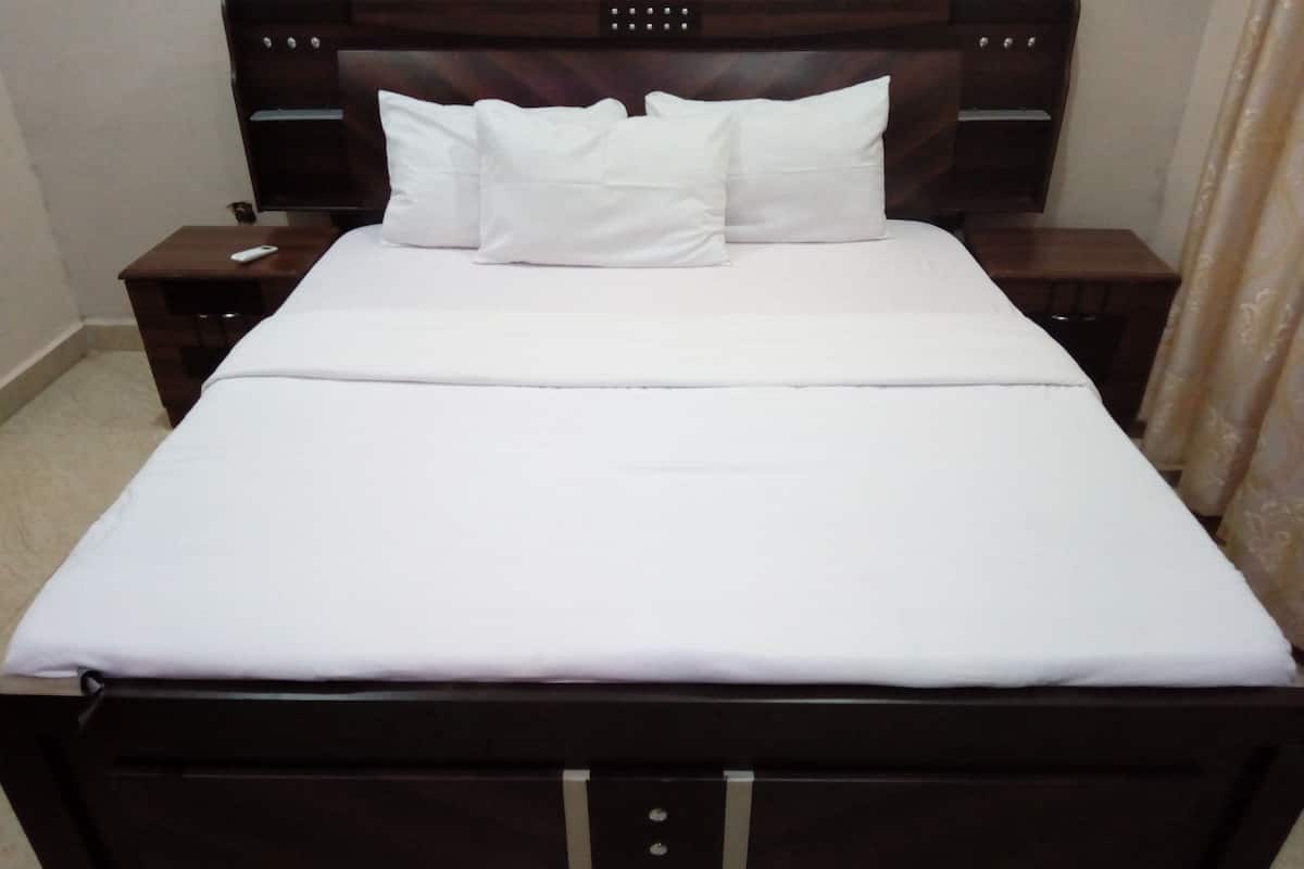 Cama bem arrumada com roupa de cama e almofadas brancas, cabeceira de madeira e mesa de cabeceira com controle remoto.