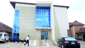 Un edificio moderno con una amplia fachada de vidrio, una entrada central con escaleras y un estacionamiento con automóviles.