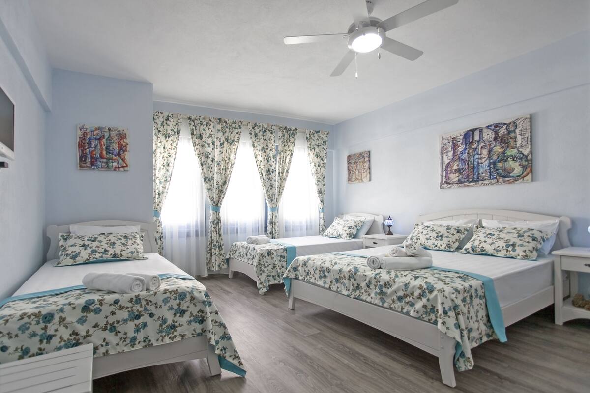 A hotel room with three single beds, floral-patterned bedding, a ceiling fan, and artwork on the walls.