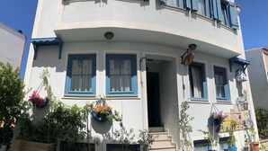 A white building with blue shutters and a curved facade. There are potted plants and hanging flower baskets outside. - Bozcaada Basak Konukevi (Bozcaada)