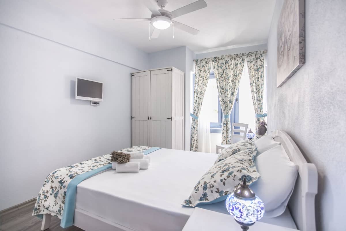 A bedroom with a bed, a ceiling fan, a TV, a wardrobe, and a window with curtains.