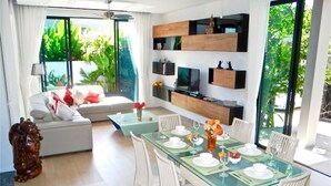 A modern dining area with a glass table, white chairs, and a TV mounted on the wall. - Baan Bua Nai Harn 3 bedrooms Villa (Rawai)