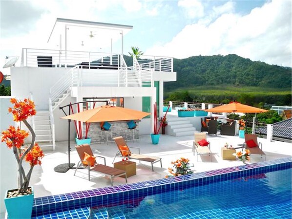 Un área de piscina moderna al aire libre con sillones de descanso y sombrillas, una escalera que lleva a un nivel superior y vistas a un paisaje verde.