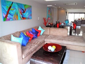 A modern living room with a beige sectional sofa, colorful cushions, abstract wall art, and a red bowl on a coffee table.