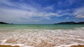 Een strand met helder turquoise water, zachte golven en heuvels in de verte.
