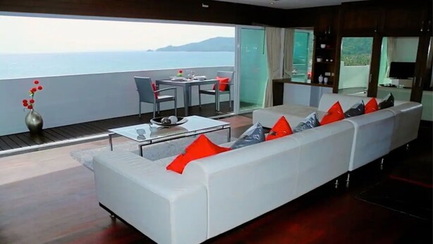 A modern living room with a white sofa, red cushions, a glass coffee table, and a view of the sea.