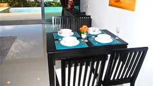 Un coin repas avec une table dressée pour quatre personnes, un tableau représentant une scène de plage et une vue sur une piscine et des montagnes.