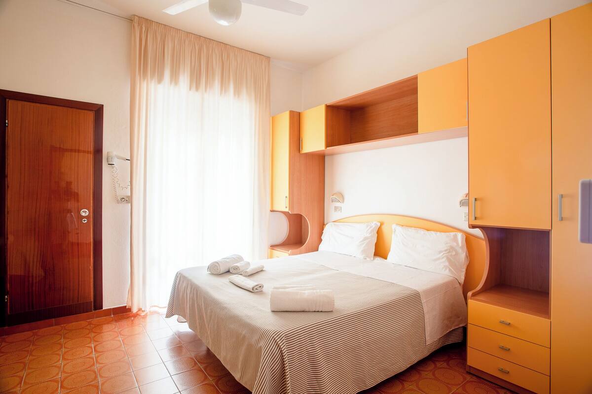A hotel room with a bed, a wooden wardrobe, a bedside table, and a window with curtains.