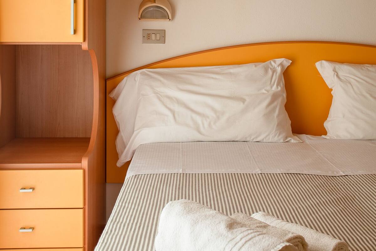 A hotel room with a bed, a nightstand, a wall-mounted light, and a towel on the bed.