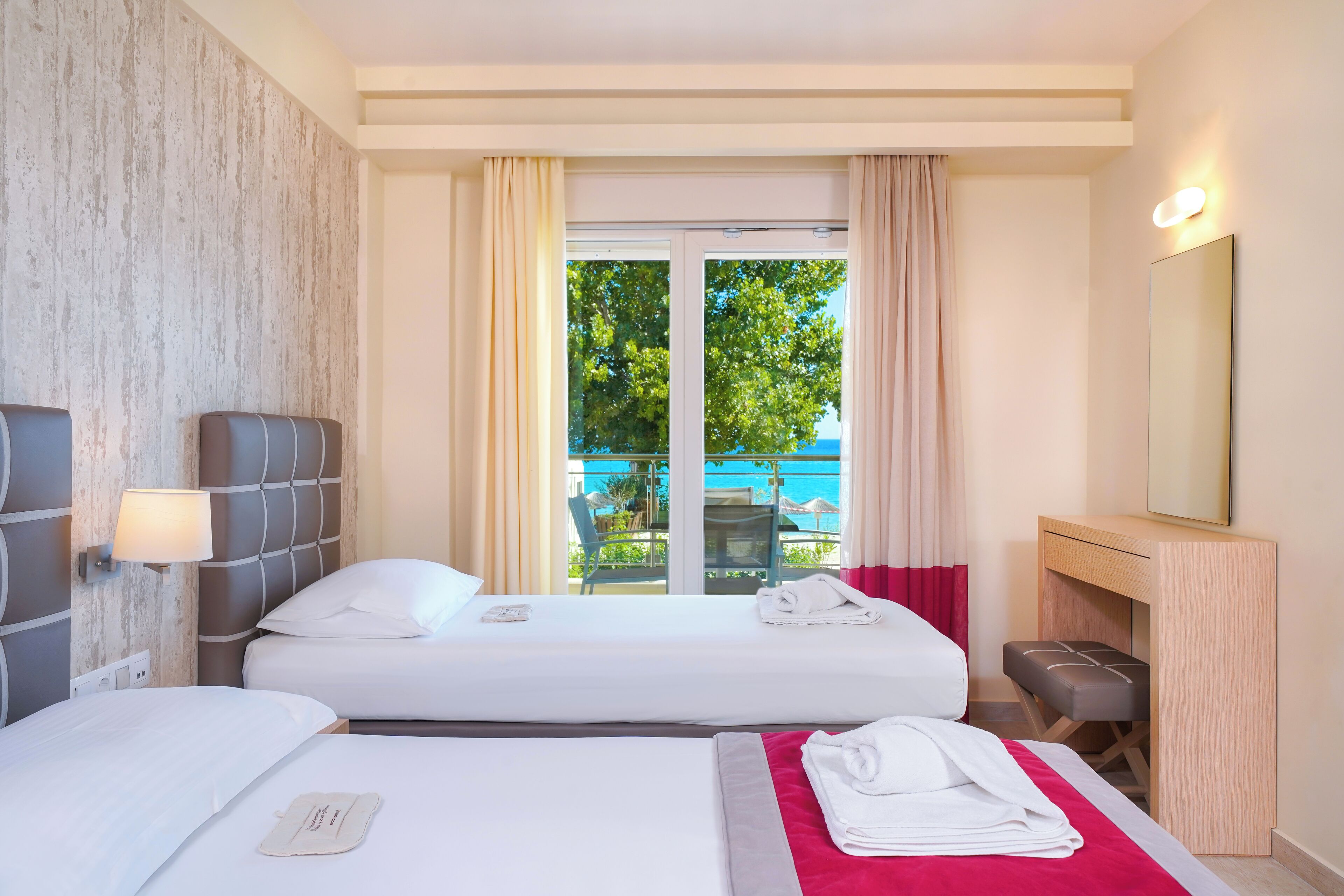 A hotel room with two beds, a desk, a chair, a mirror, and a view of the outdoors.