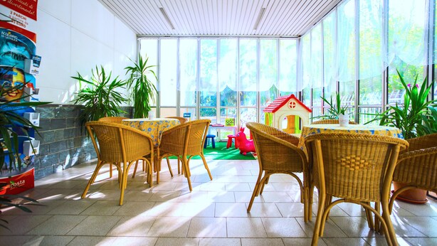 Un espacio interior luminoso con mobiliario de mimbre, una zona de juegos con una casita y ventanas amplias.