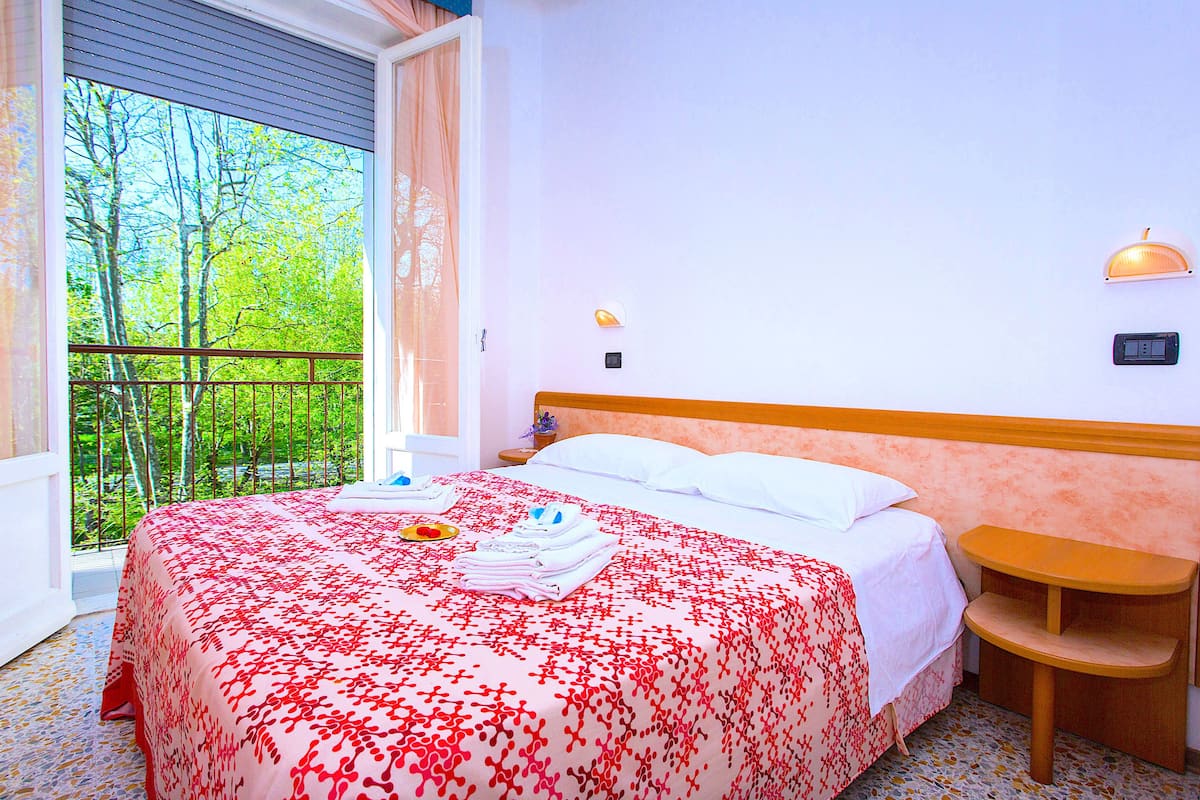 A hotel room with a bed, a nightstand, a wall-mounted lamp, and a window with a view of trees.