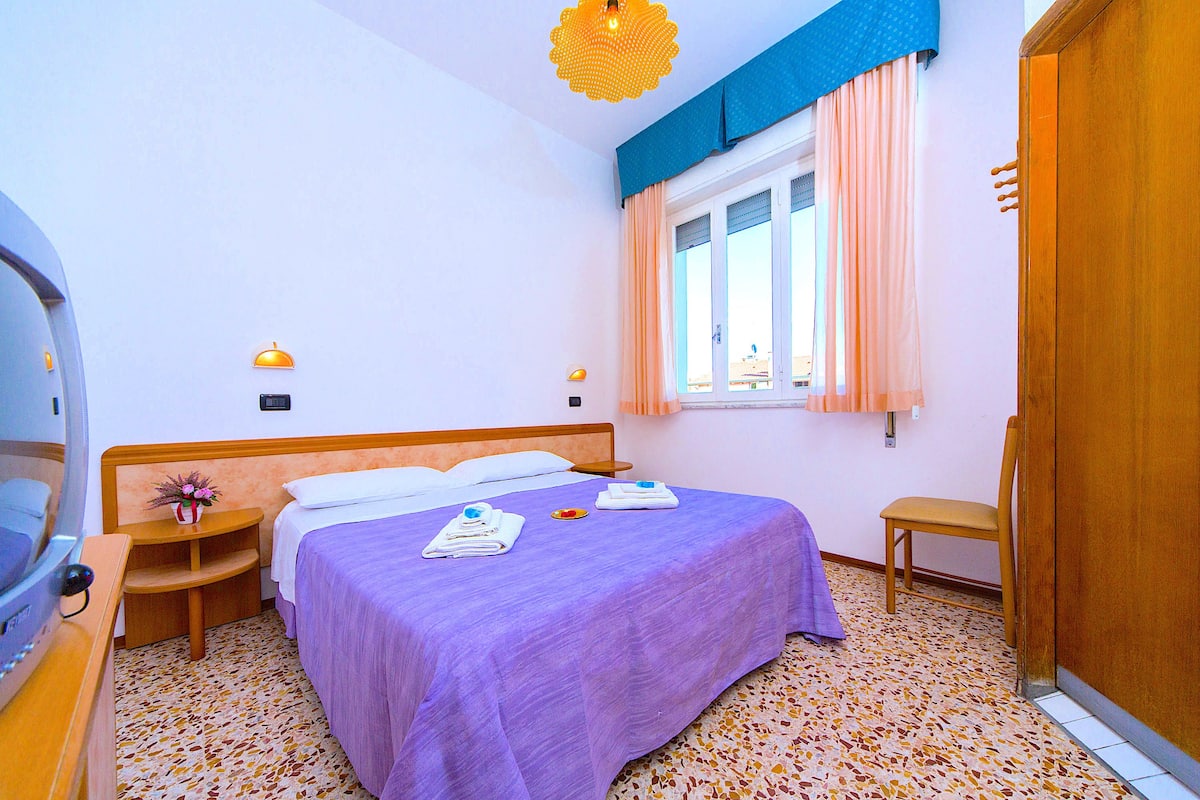 A hotel room with a bed, bedside tables, a chair, a wardrobe, a window with curtains, and a ceiling light fixture.