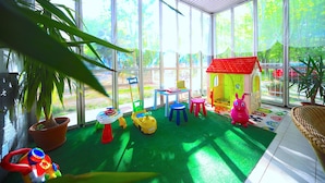 Un espacio de juego interior luminoso, con suelo verde, una casita roja y diversos juguetes.