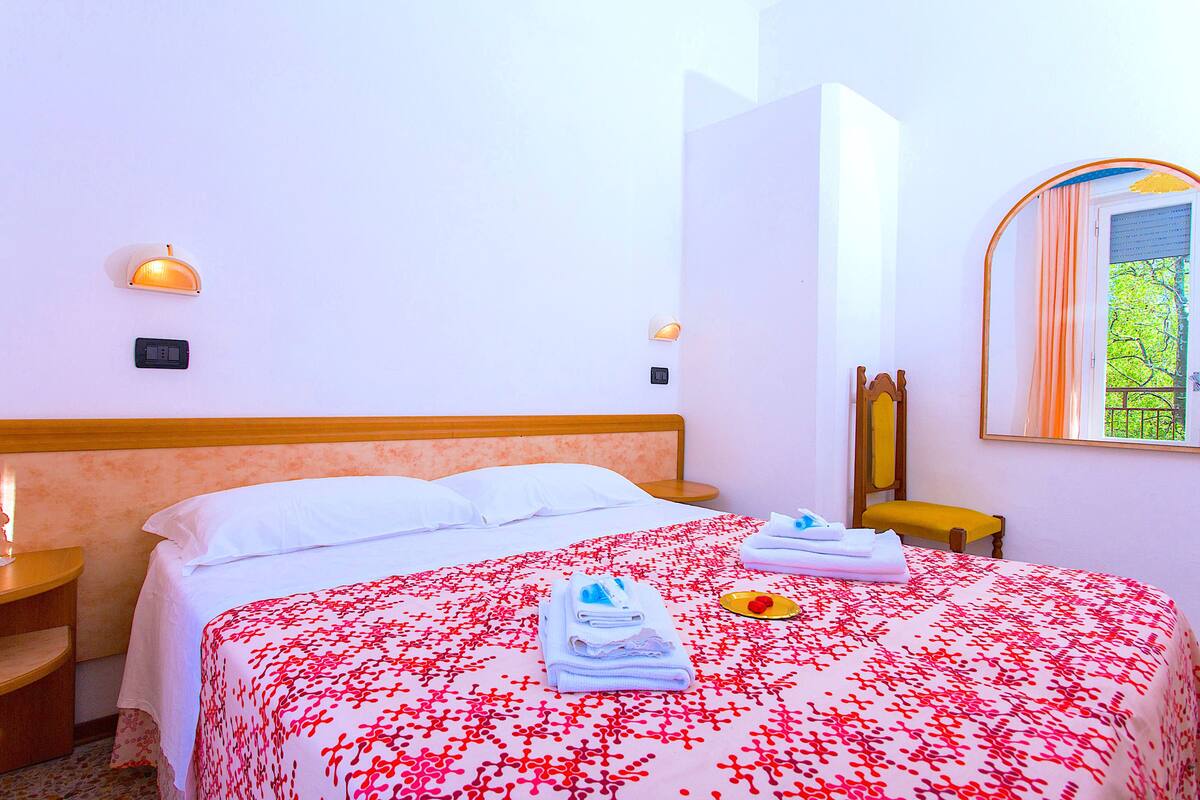 A hotel room with a bed, a nightstand, a mirror, and a window with a view of greenery.