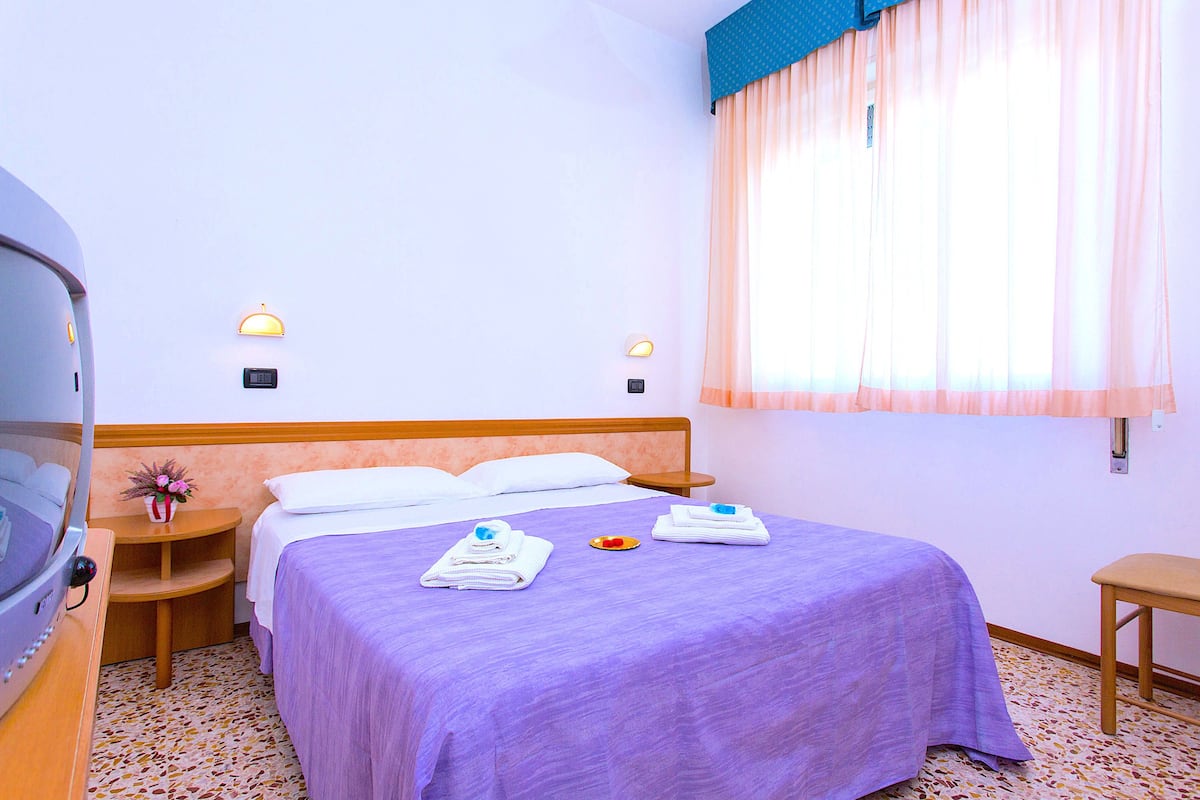 A hotel room with a bed, two bedside tables, a small plant, and a window with curtains.