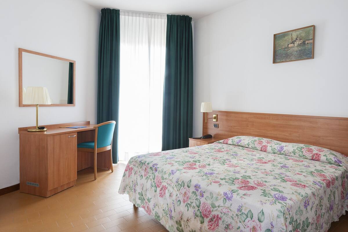 A hotel room with a bed, desk, chair, and a painting on the wall.