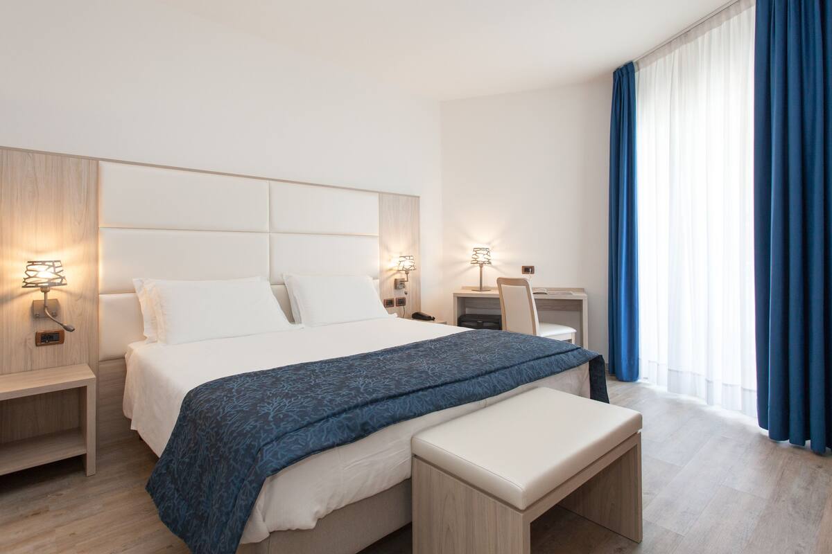 A modern hotel room with a large bed, bedside tables, a desk, and a window with curtains.