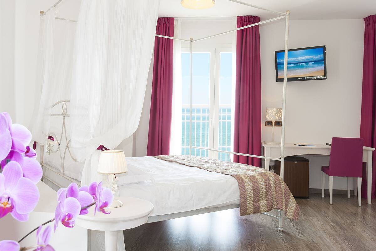 A hotel room with a four-poster bed, a desk, a chair, a TV, and a window with curtains.