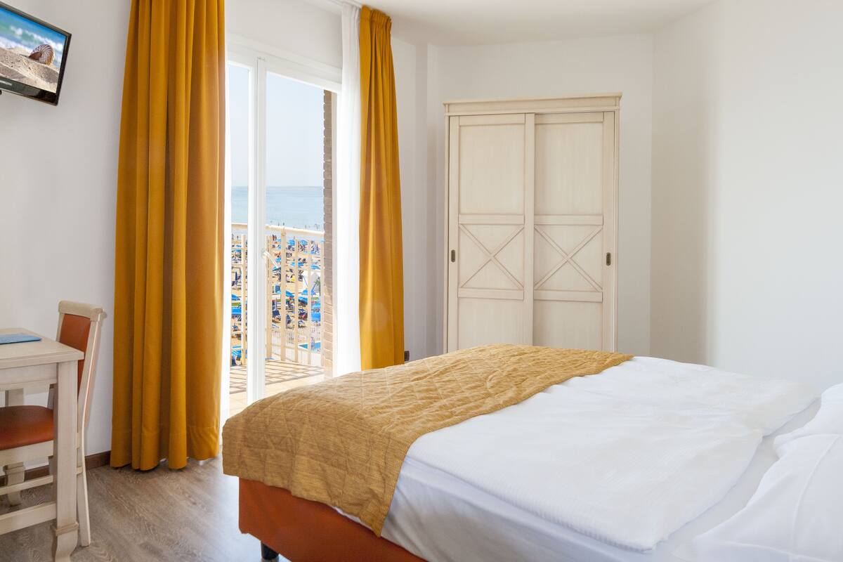 A hotel room with a bed, a desk, and a chair. There is a window with yellow curtains and a view of a beach and buildings.