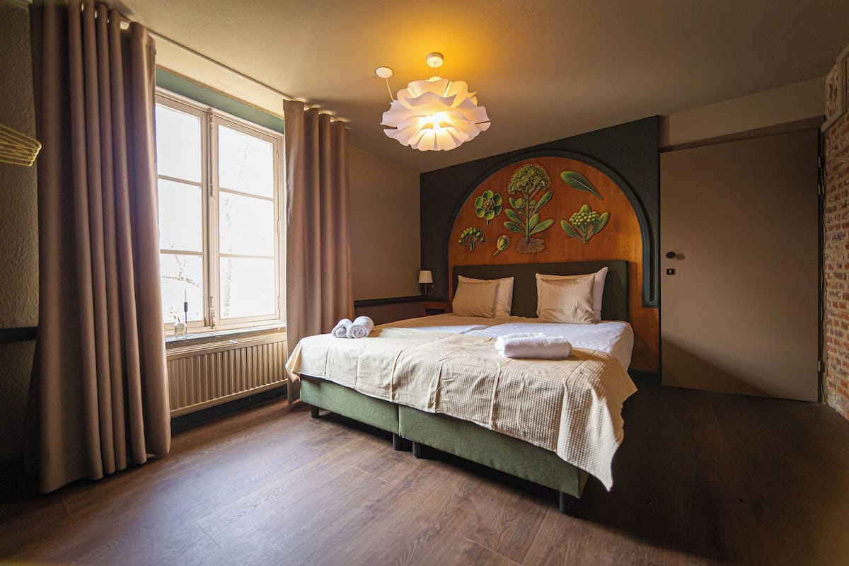Un lit bien fait, avec une tête de lit ornée de motifs floraux, une fenêtre avec des rideaux et un radiateur placé sous la fenêtre.