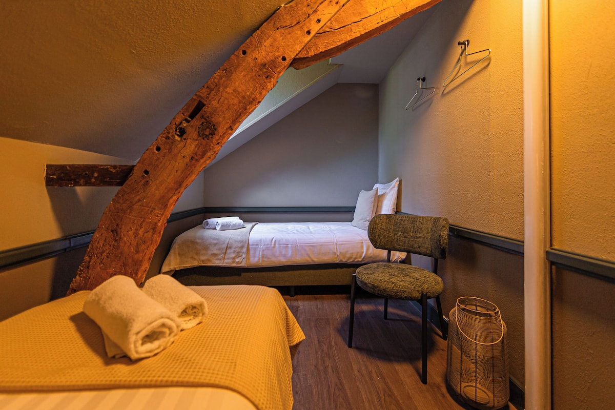 Une chambre confortable avec un plafond en pente, un lit sur lequel est posée une serviette enroulée, une chaise et un panier en osier.