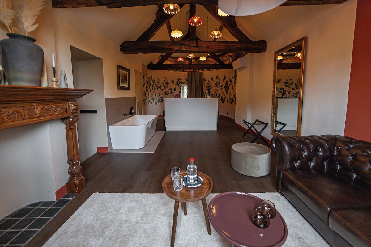 Un vaste espace de vie avec un plafond en bois, un canapé en cuir marron, une petite table ronde avec un verre et une bouteille, et une baignoire blanche en arrière-plan.