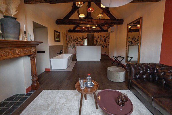 A spacious living area with a wooden ceiling, a brown leather sofa, a small round table with a glass and a bottle, and a white bathtub in the background.