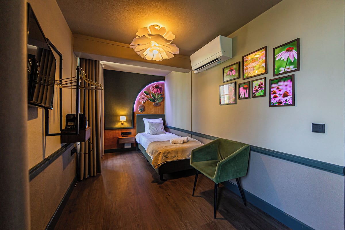 Une chambre d’hôtel comprenant un lit, un bureau, une chaise, un miroir, un climatiseur fixé au mur et des tableaux représentant des motifs floraux.