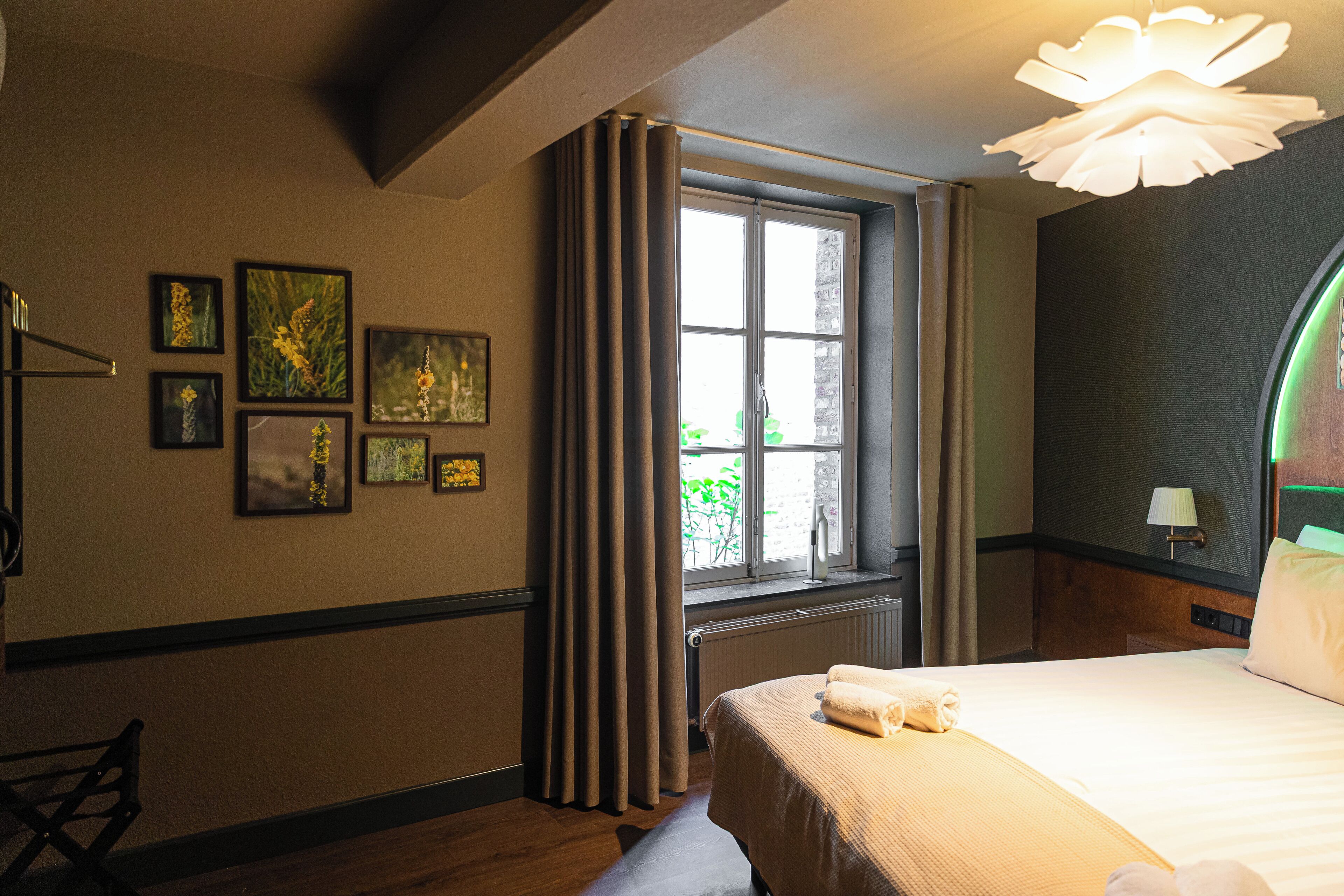 Une chambre d’hôtel avec un lit, une fenêtre avec des rideaux, un radiateur et des tableaux encadrés au mur.