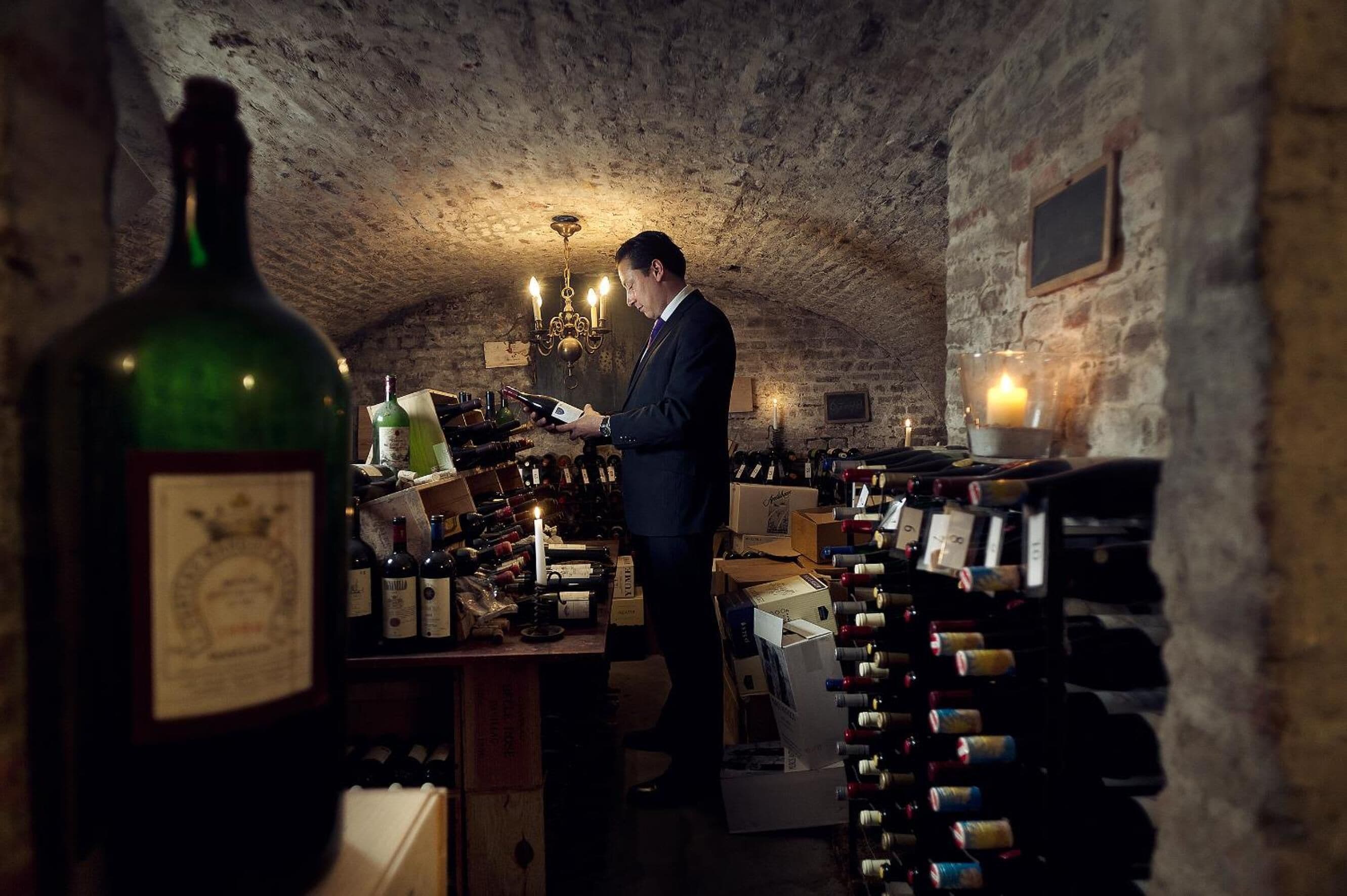 Un hombre con traje en un sótano tenuemente iluminado, con numerosas botellas y cajas de vino.