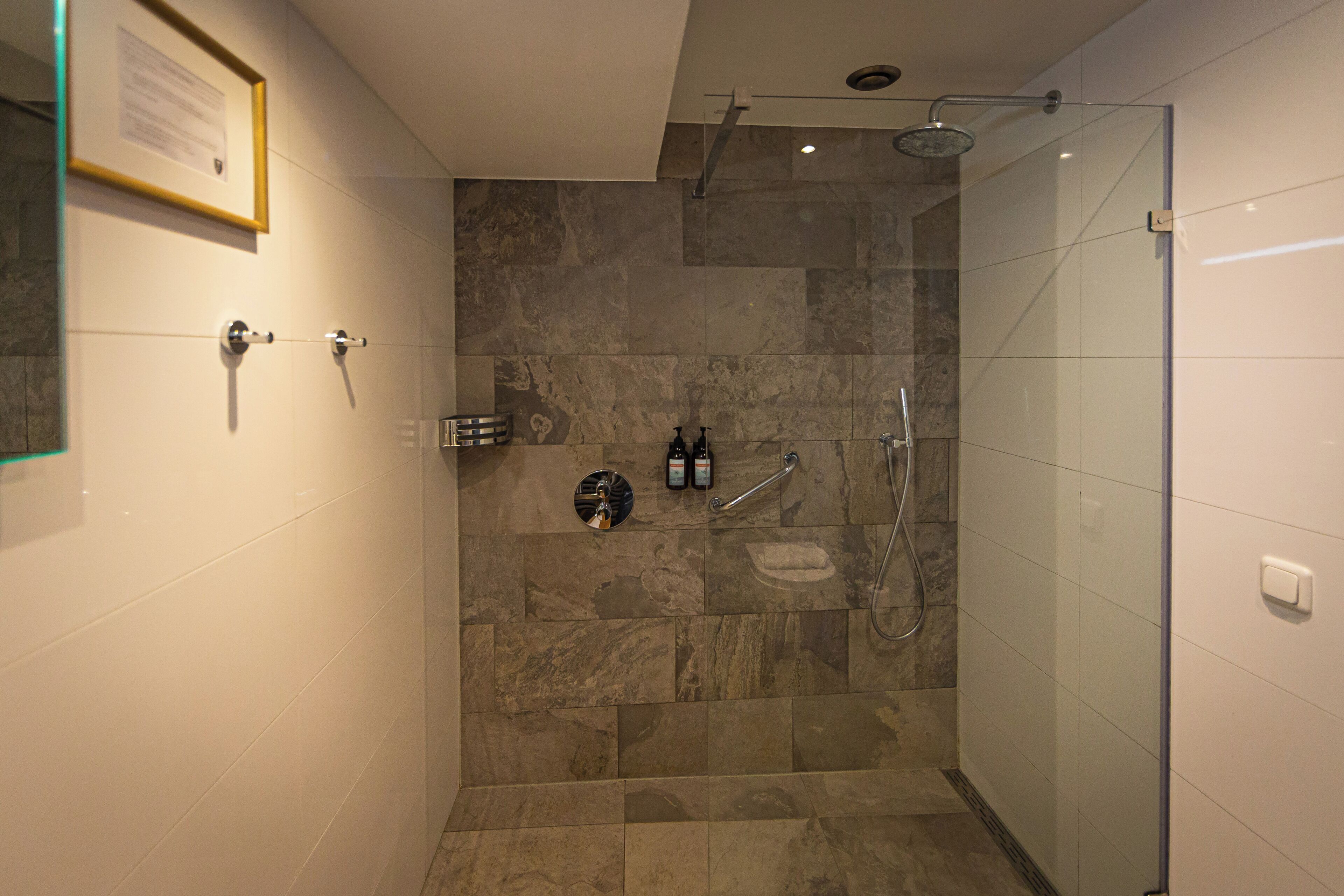 Un baño moderno con cabina de ducha de vidrio, zona de ducha con azulejos de mármol y lavabo de pared con dispensador de jabón.