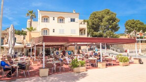 An outdoor café with tables and chairs, a covered seating area, and a building with multiple windows and balconies in the background. - Apartamentos Niza (Calvia)