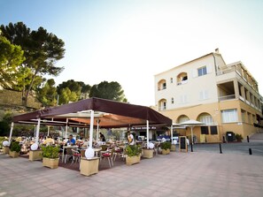 An outdoor cafe with tables and chairs, a building with balconies, and a stone wall in the background. - Apartamentos Niza (Calvia)