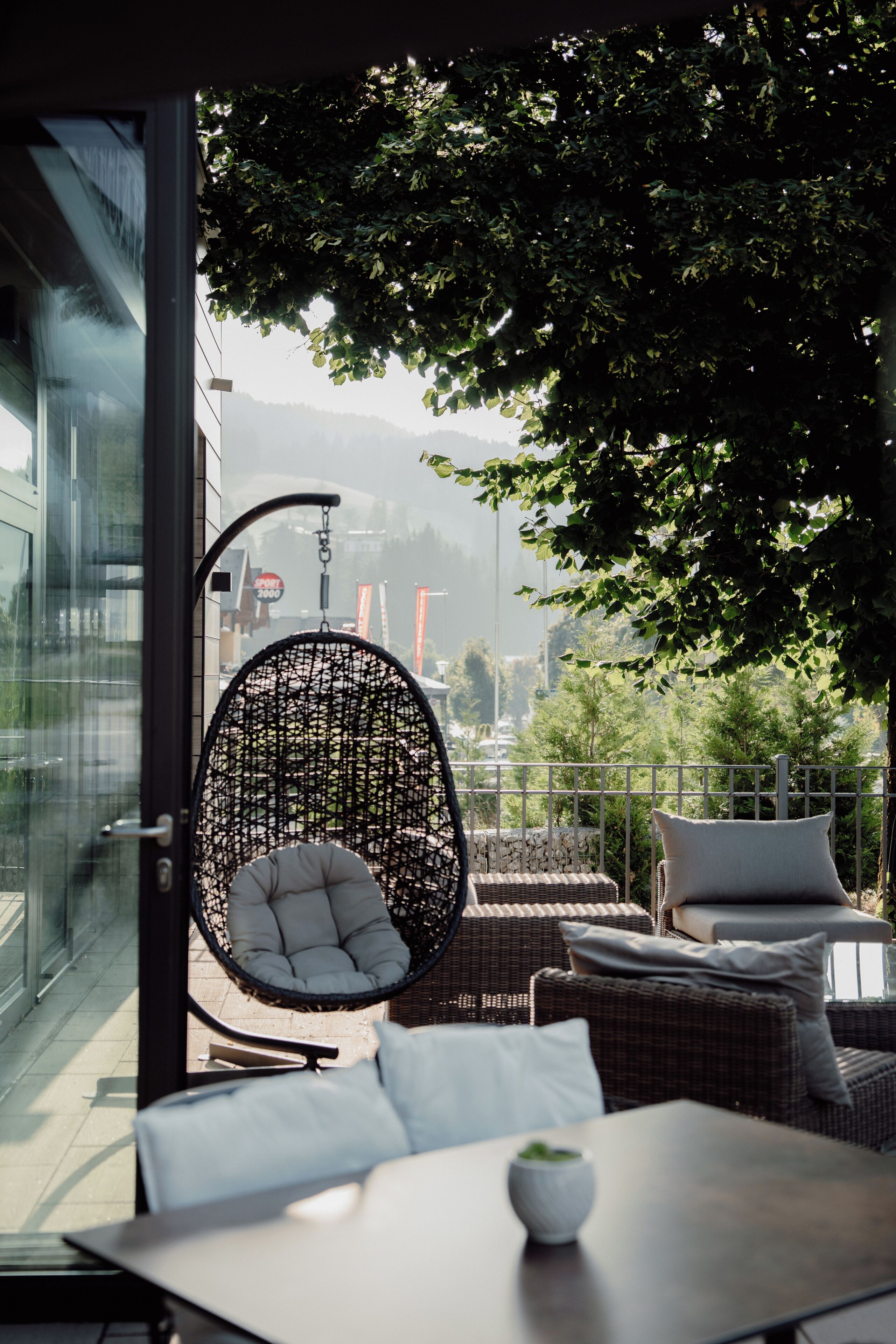A modern outdoor seating area with a wicker chair and cushions, a table with a white bowl, and a view of a cityscape through a glass wall.