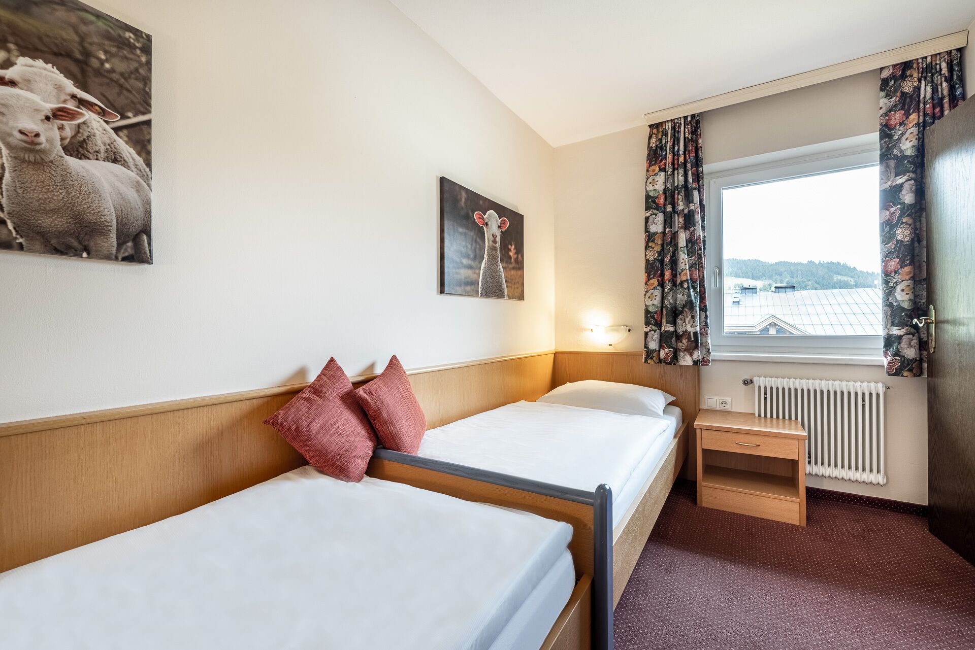 Hotelkamer met twee enkele bedden, een nachtkastje, een lamp, een raam met bloemen gordijnen en uitzicht op gebouwen.