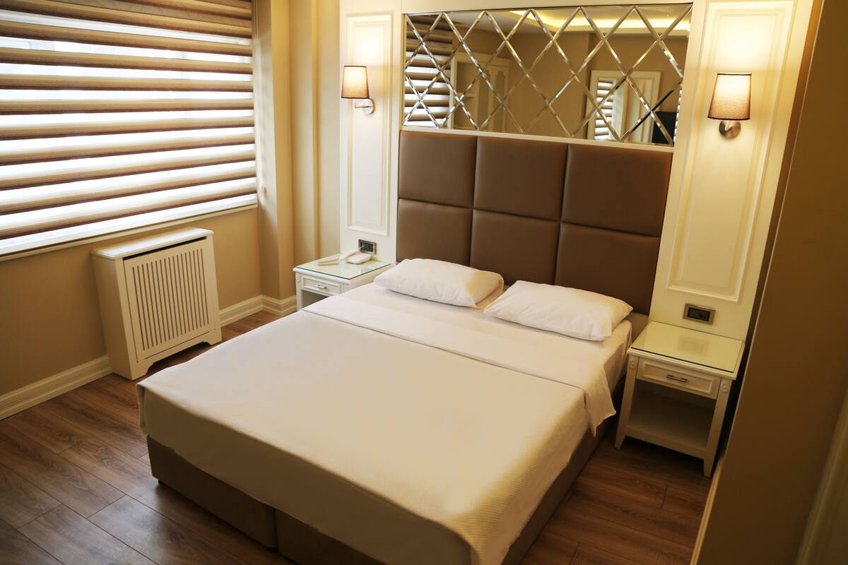A neatly made bed in a hotel room with a large mirror above it, a bedside table with a lamp, and a radiator under the window blinds.