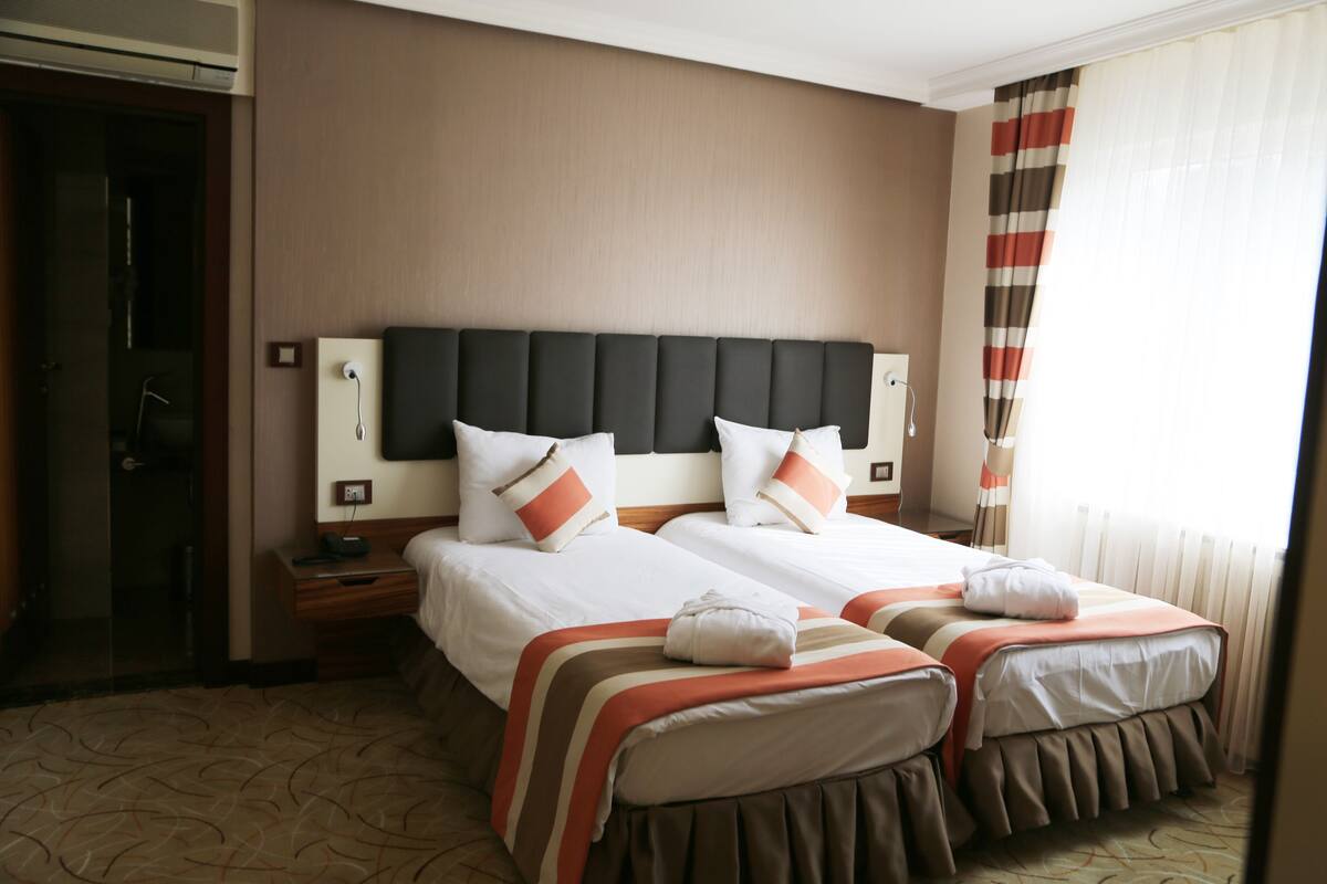 A hotel room with two beds, a headboard, bedside tables, and a window with curtains.